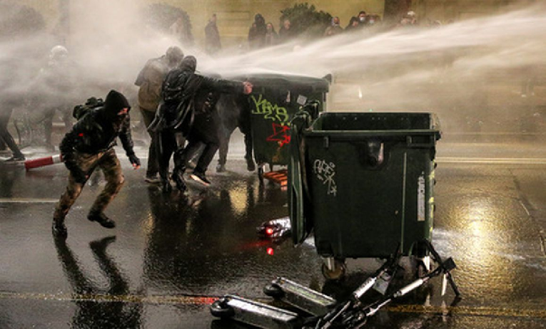 Протестующие укрываются за мусорными баками от струи водомета. Фото Ираклий Геденидзе/Reuters/Scanpix/LETA