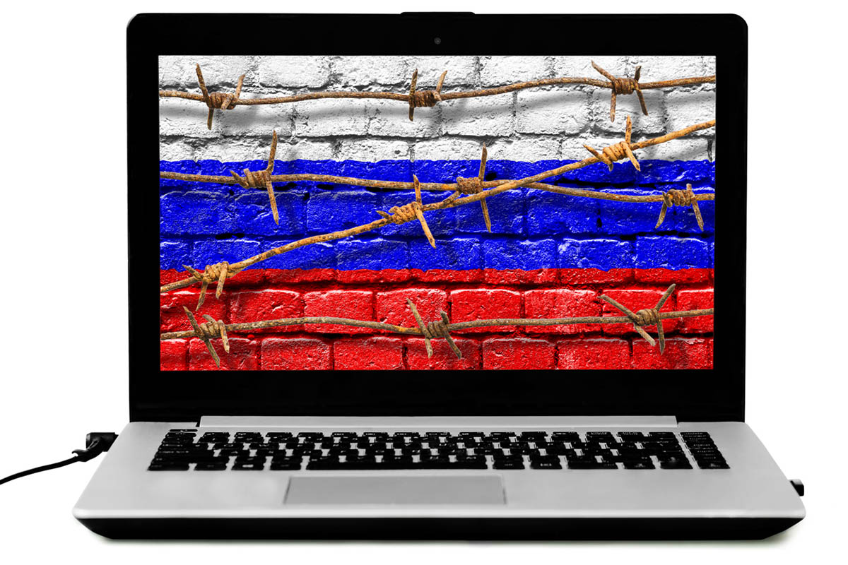 Экран ноутбука с колючей проволокой и российским флагом на кирпичной стене. Фото Dzurag/istockphoto